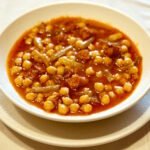 Primer plano de un guiso de garbanzos con trozos de chistorra roja, virutas de jamón serrano y tallos de borraja verde en un plato de barro artesanal